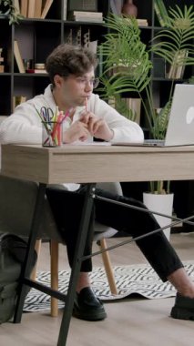 Odaklanmış adam dik oturur ve dizüstü bilgisayar izlerken kalemi ağzının yanına yerleştirir. Ofis ayarları kitap rafı ve canlı bitkilerle dolu bir masa içerir