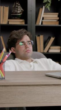 Adam koltuğa uzanıp dizüstü bilgisayarını rahatça izliyor. Kompozisyon masa ögeleri ve arkaplan dolusu kitap ve yeşillik içerir