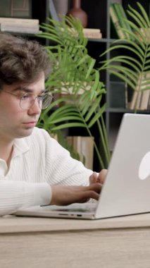 Gözlüklü öğrenci, masa başında kitaplar ve bitkilerle birlikte dizüstü bilgisayarda yazmaya odaklanıyor. İç mekan üretken öğrenme ortamı yaratır