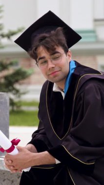 Yüzünde hüzün belirtileriyle mezun olmuş, bankta oturmuş gülümsüyor. Adam diplomasını sarmış ve üniversite kampüs parkına bakıyor.