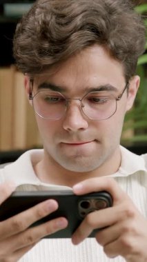 Gözlüklü genç adam telefonda video oyunlarını sever. Ofisteki sandalyede rahatça oturur. Brunet etrafı kitaplar ve bitkilerle çevrili.