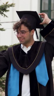 Mezun adam akademik cübbeyle selfie çekerken gülümsüyor. Öğrenci, yüksek binanın önünde duran havan tahtasına hafifçe uyum sağlıyor.