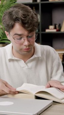 Odaklanmış üniversite öğrencisi final sınavlarına hazırlanırken ders kitabı okur. Adam, hafif ev kütüphanesinde dizüstü bilgisayar ve defterlerle masada oturuyor.