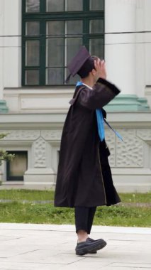 Yüksek lisans öğrencisi şapkasını çıkararak üniversite binasına doğru dönüyor. Adam büyük tarihi binaya neşeyle el sallıyor.