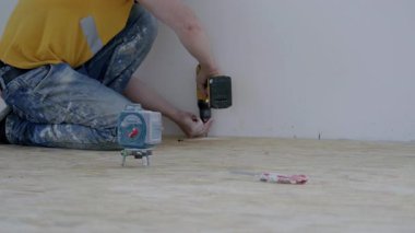 Erkek inşaat işçisi yerde diz çökerek, materyalleri sabitlemek için matkap kullanıyor, alet ve çalışma alanı elementleri ile duvarcılık ve sıva becerilerini gösteriyor