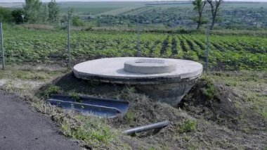 Yeşillik ve tarım alanlarıyla çevrili kırsal bir ortamda bulunan su pompası ve depolama ünitesi, kamu suyu erişimi için hayati kaynakları vurguluyor.