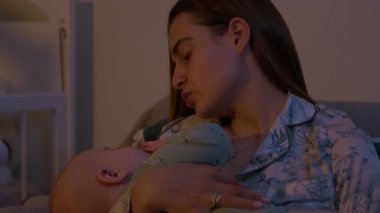 Huzurlu genç anne, geceleri kucağında bebek beşiği taşıyor. Pijamalı sevgi dolu kadın, akşam yatak odasında oturan küçük çocuğu sakinleştirir.