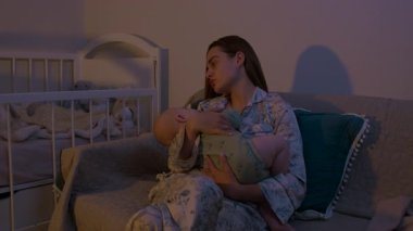 Pijamalı genç anne kanepede oturan beşikte uyuyan bebeği nazikçe kucaklar. Kadın, çocuğu yatak odasında rahat ettiriyor.