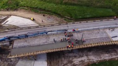 İşçiler aktif olarak yenileme altındaki bir köprüye beton döküyor, açık hava ortamında işbirliği ve inşaat tekniklerini vurguluyor