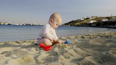 Genç çocuk kumsaldaki yumuşak kumlarda oynuyor. Etrafı renkli oyuncaklarla çevrili. Hafif dalgalar kıyıya vuruyor.