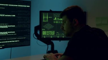 Veri analizi yapan, kod ve grafikle çevrili birçok ekranla çevrili bir erkek profesyonel, yüksek teknoloji ortamı ve çalışmalara yoğunlaşmayı gösteriyor.