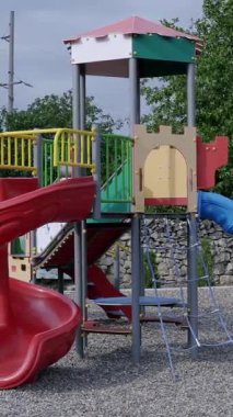 Kırmızı kaydırak ve mavi rampalı canlı oyun alanı ekipmanları, arka planda ağaçlar olan çakıllı bir alana kurulmuş, dışarıda oyun ortamı yaratıyor.