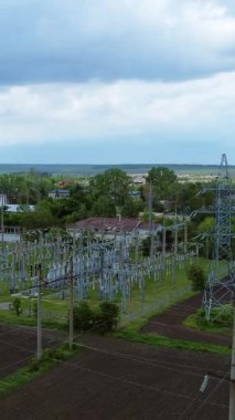 Yüksek voltajlı elektrik hatları ve santraller, yeşilliklerle çevrili geniş bir arazide ve dramatik bulutlu bir gökyüzünün altında, enerji taşıma sistemlerini gösteriyor.