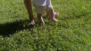 Bebek, yetişkinlerin desteğiyle yumuşak çimenlerde yürümeyi öğreniyor, doğadaki çocukluk keşiflerinin heyecanını ve masumiyetini vurguluyor.