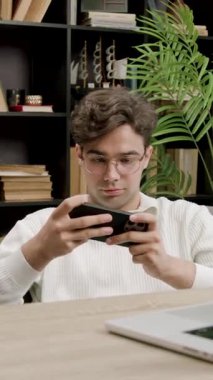 Genç adam cep telefonuyla bilgisayar ve kitaplarla ofis masasında oyun oynuyor. Sosyal medyada vakit harcamayı erteliyor