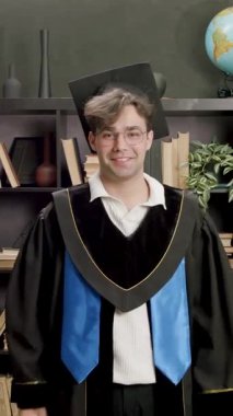 Mezuniyet elbisesi ve havan tahtasındaki genç adam diplomasını gururla taşıyor. Öğrenci başarıyı kollarını kaldırıp kütüphanede gülümseyerek kutluyor.