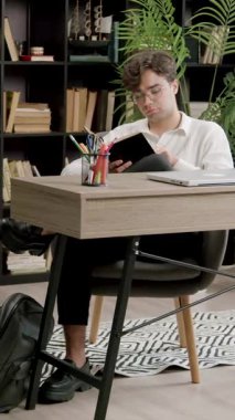 Genç bir erkek öğrenci kütüphanede masa başında oturmuş kitap okuyor ve ders çalışıyor. Brunet masadaki dizüstü bilgisayarları ve defterleri kullanır