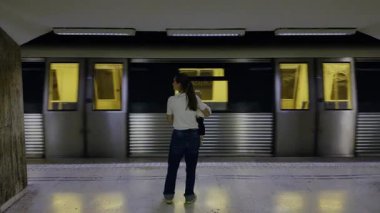 Kadın banliyö metro istasyonunda duruyor, trenin hareket bulanıklığı ile arka planda, şehir seyahatinin ve günlük yaşamın özünü yakalıyor.