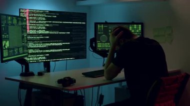 Masasında kod ile dolu birden fazla ekranı olan, karanlık, teknolojik odaklı çalışma alanında stres ve hüsran sergileyen, dijital görevlerin zorluklarını vurgulayan kişi