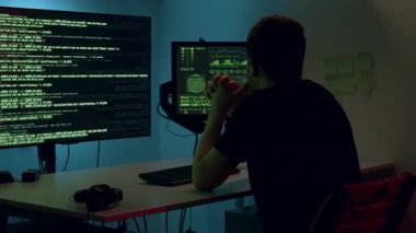 Erkek yazılım geliştirici karanlık bir ortamda çift monitörlerdeki kodları analiz ediyor. Programlamanın karmaşıklığını ve teknoloji odaklı çalışma alanını gösteriyor.