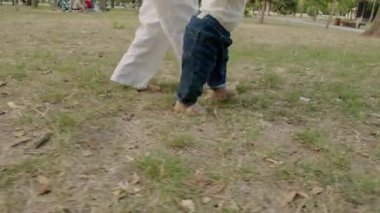 Bebek mutlu bir şekilde çimenli zeminde ilk adımlarını atar, yetişkin desteğiyle ilerler, erken yürüyüşün heyecanını yakalar ve cesaret bağını yakalar.