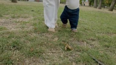 Bebek, çimenli bir parkta annesinin desteğiyle ilk adımlarını atıyor. Yeni hareketin heyecanını ve ebeveynler ile çocuklar arasındaki bağları vurguluyor.