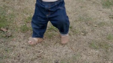 Bebek yumuşak çimenlerde yürümeye başlar, kot pantolon ve sandalet giyer, ilk adımların heyecanını yakalar ve açık hava keşiflerinin güzelliğini yakalar.