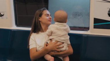 Kadın metroda bebeğini kucaklıyor, ikisi de pencereden dışarı bakıyor, şehir çevresi ve hayat yolculuğu arasında samimi bir an yaşıyor.