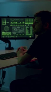 Erkek girişimci, karanlık bir odada kod ve analizlerle dolu çift taraflı bilgisayar monitörleriyle, dijital yenilik ve konsantrasyon atmosferini vurguluyor.