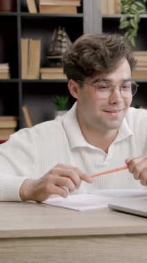 Adam internette konuştuktan sonra gülümser ve sandalyeye yaslanır. Masada kitapların düzenli arkaplanlı dizüstü bilgisayar ve kırtasiye malzemeleri var