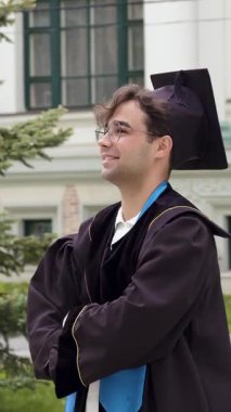 Gururlu mezun kolları bağlı ve dik dik bakıyor. Akademik giysili öğrenci kutlamalardan önce bina yakınında poz veriyor