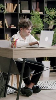 Odaklanmış öğrenci, arka planda sırt çantası ve kitaplıklarla ev ofisinden ders çalışırken dizüstü bilgisayar kullanıyor. Adam doğal pencere ışığından hoşlanıyor.