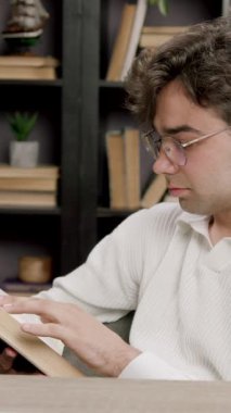 Genç üniversite öğrencisi hafif ev kütüphanesinde ders kitabı okuyor. İnsan, kitaplarla çevrili rahat ve sessiz bir ortamda çalışmalara odaklanır.