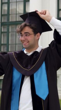 Heyecanlı mezun adam havan topunu eğip telefon açısını değiştiriyor. Önlüklü öğrenci, ladin ağacıyla klasik binanın yakınında poz veriyor