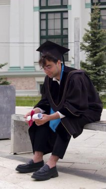 Akademik önlük ve kep giyen genç erkek mezun kampüsün yanındaki bankta diplomasını mutlu bir şekilde tutuyor. Adam gelecekte işlerinde başarılı olmayı hayal ediyor.