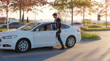 Erkek alıcı, açık kapının yanında duran beyaz bir sedan 'ın durumunu kontrol ediyor. Sıcak güneş ışığı ve yeşillik hoş bir atmosfer yaratıyor.
