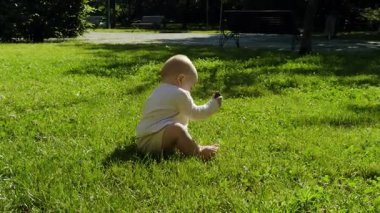 Güneşli bir parkta yemyeşil çimlerin üzerinde oturan bebek, doğa ve yumuşak güneş ışığıyla çevrili, merakla etrafı keşfediyor, bir merak hissi uyandırıyor.