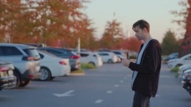 Sıradan genç bir adam park alanında yürüyor, park halindeki araçlarla çevrili akıllı telefonu kontrol ediyor, işe hazırlanıyor.