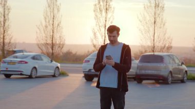 Erkek bireyler günbatımında park alanında akıllı telefonla etkileşime girerken, arka planda park edilmiş araçlar çağdaş araba paylaşım deneyimini göstermektedir.