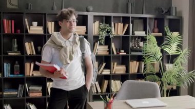 Sırtında sırt çantasıyla şık ve yakışıklı bir öğrenci ve kitapları odaya bakıyor ve gülümsüyor. Esmer çocuk prestijli bir eğitim okulunun modern sınıfında.