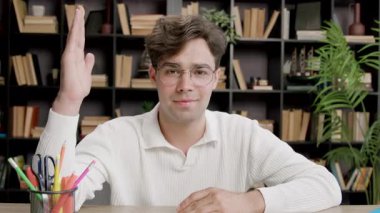 Bankta oturan bir erkek öğrenci doğrudan kameraya bakıyor, alaycı bir şekilde gülümsüyor ve cevabını vermek, fikrini söylemek için elini kaldırıyor. Terbiyeli.