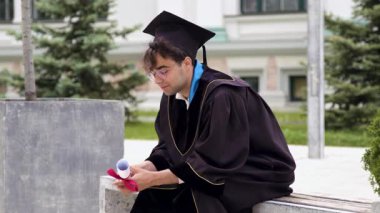 Mezuniyet elbiseli, siyah cüppeli ve şapkalı üniversite kampüsünün avlusunda bir sandalyeye oturmuş mezuniyet diplomasını gururla elinde tutuyordu. Uluslararası Çalışmalar