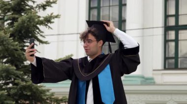 Şapkalı ve cüppeli modern mezun çocuk. Genç adam, üniversiteden başarılı bir şekilde mezun olan öğrencilerin mezuniyet töreninden birkaç dakika sonra aileleri ve arkadaşlarıyla konuşuyor.