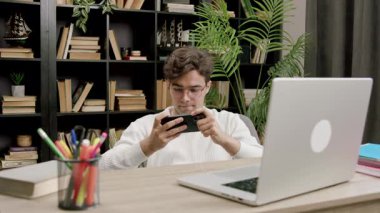 Okulun kampüs kütüphanesinde akıllı telefondan online spor videoları izleyen şaşkın, heyecanlı çocuk. Üniversite bankında elinde telefonla çevrimiçi mobil oyunlarda erkek kazanan