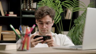 Okulun kampüs kütüphanesinde akıllı telefondan online spor videoları izleyen şaşkın, heyecanlı çocuk. Üniversite bankında elinde telefonla çevrimiçi mobil oyunlarda erkek kazanan