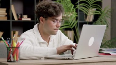 Genç bilişim uzmanı dikkatle yazıyor ve dizüstü bilgisayardaki klavyeye odaklanmış durumda. Gözlüklü çocuk modern bir ofiste çalışıyor ve web siteleri kuruyor. İş yerindeki sevimli çocuk.