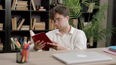 Ciddi bir öğrenci kitap okuyor ve üniversite projesi için fikirler arıyor. Kütüphanede dizüstü bilgisayar kullanarak araştırma çalışması, sınıf raporu üzerinde çalışmak. Gözlüklü çocuk derslerine ve öğrenmeye odaklanmış.