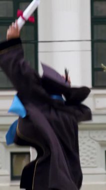 Happy and excited graduate boy in graduation gown and cap with graduation diploma in hand jumps up and dances. Young man with glasses successfully graduated from higher education
