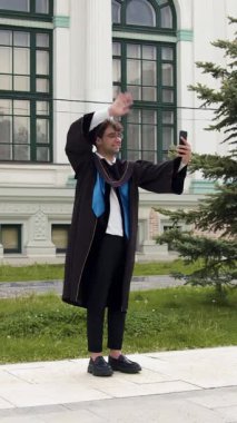 Şapka ve siyah bornoz giyen modern mezun telefonda selamlaşır. Genç adam üniversitenin mezuniyet töreninden anları göstermek için aileleri ve arkadaşlarıyla internette sohbet ediyor.