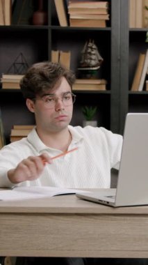 Angry boy with glasses, nervous about the large volume of lessons and projects for college. Student preparing for lessons and exams at home in a modern office with a library in the background